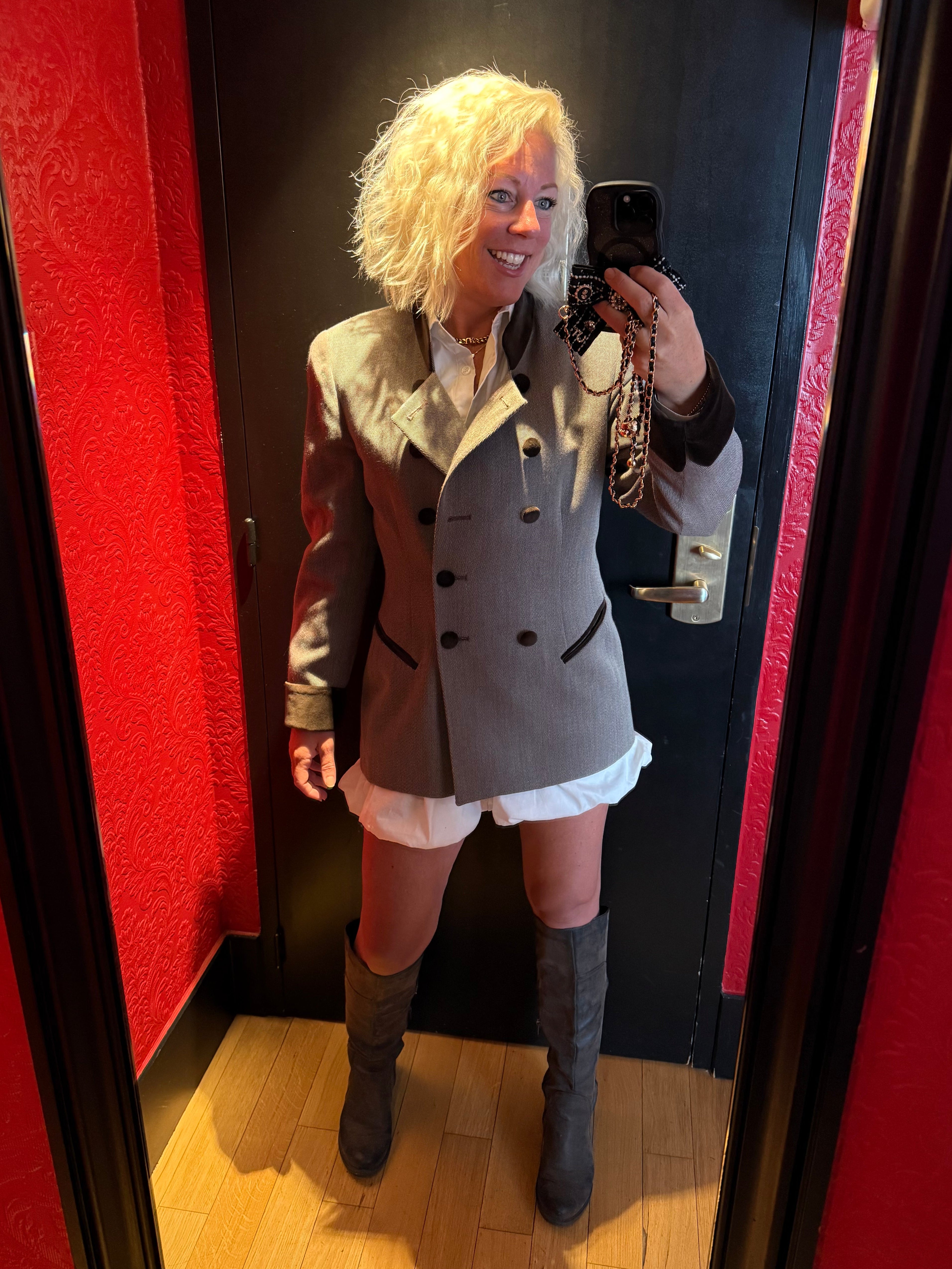 Person taking a mirror selfie wearing a coat and skirt in a room with red carpet and wooden floor.