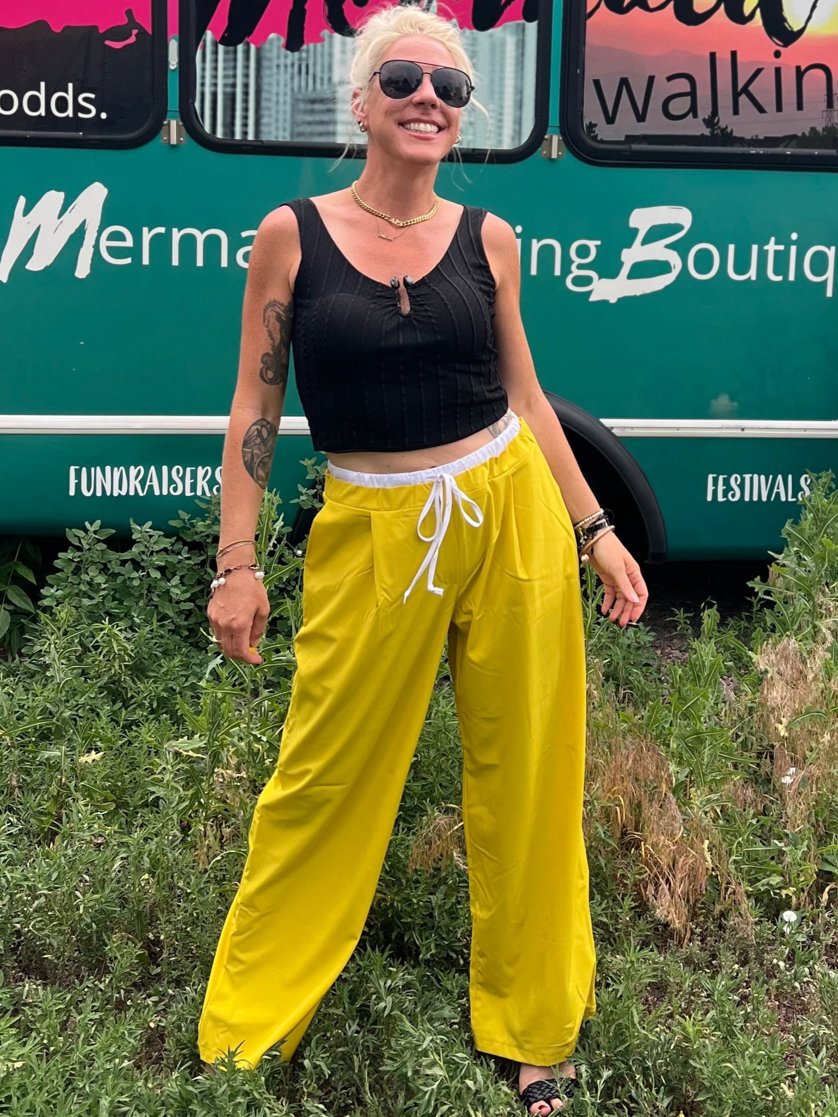 Woman in yellow pants and black top standing in front of a mobile boutique bus.