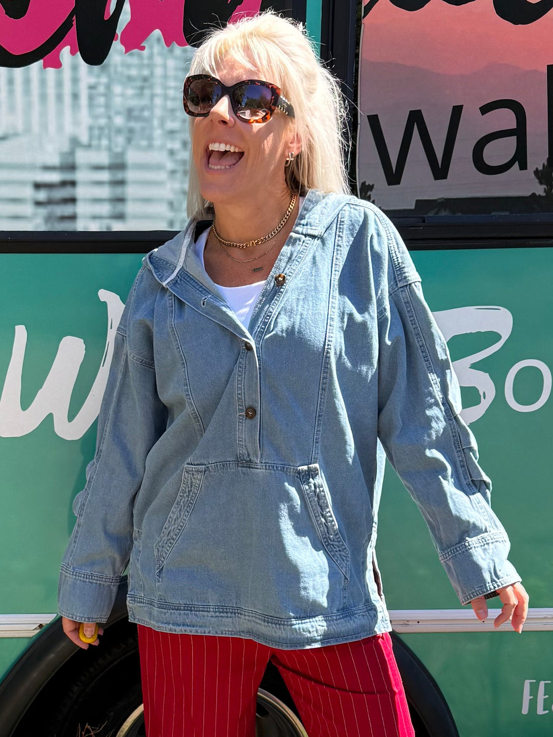A woman wearing a light blue denim jacket and bright red pants stands in front of a teal bus with 'Mermaid Walking Boutique' printed on the side.