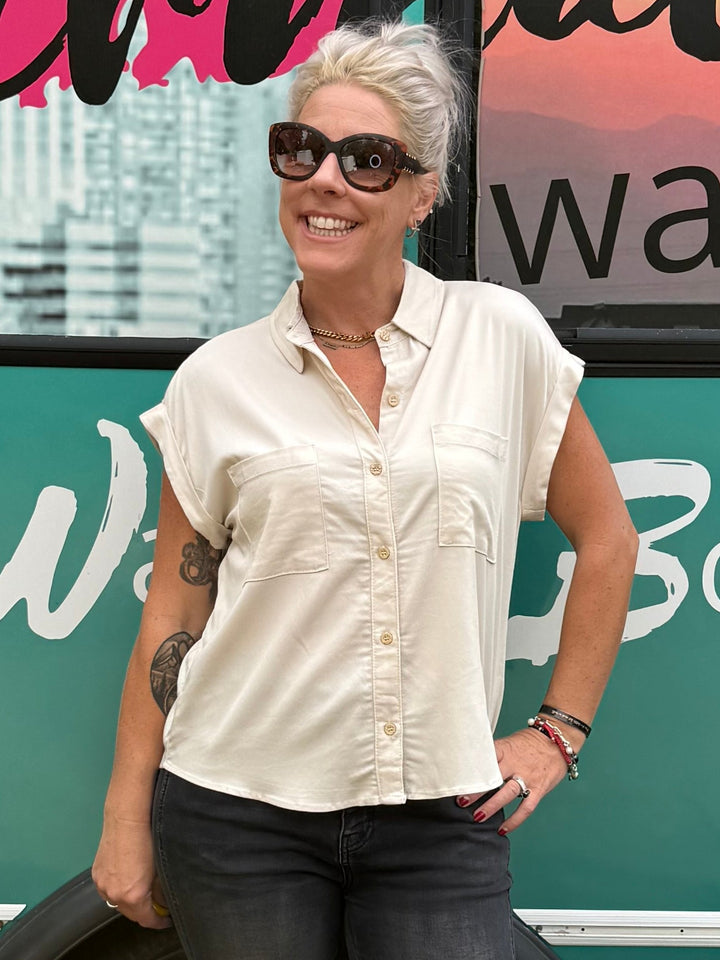 A woman wearing a cream short-sleeve button-up shirt, dark jeans, and sunglasses stands in front of a teal bus with &