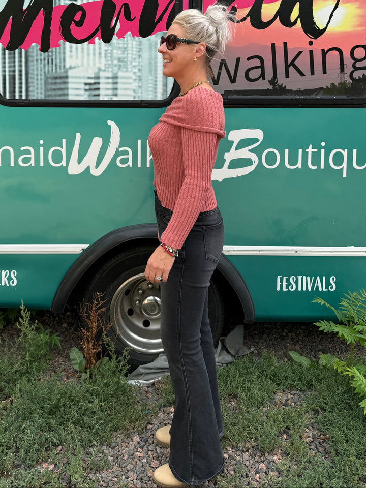 A woman wearing a pink ribbed long-sleeve top and black flared pants stands in front of a teal bus with &