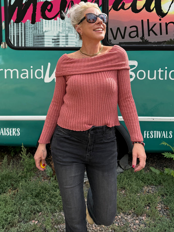 A woman wearing a dusty rose ribbed off-the-shoulder sweater and black jeans stands in front of a teal bus with &