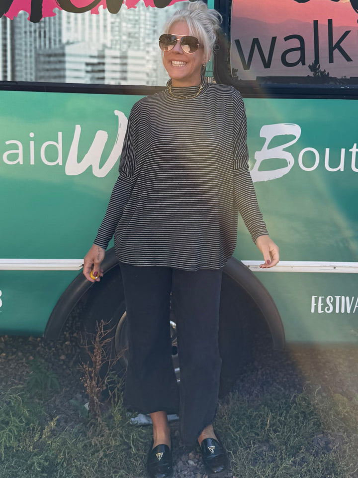 A woman wearing a dark gray long-sleeve mock neck top, black pants, and black loafers stands in front of a turquoise and green bus with &