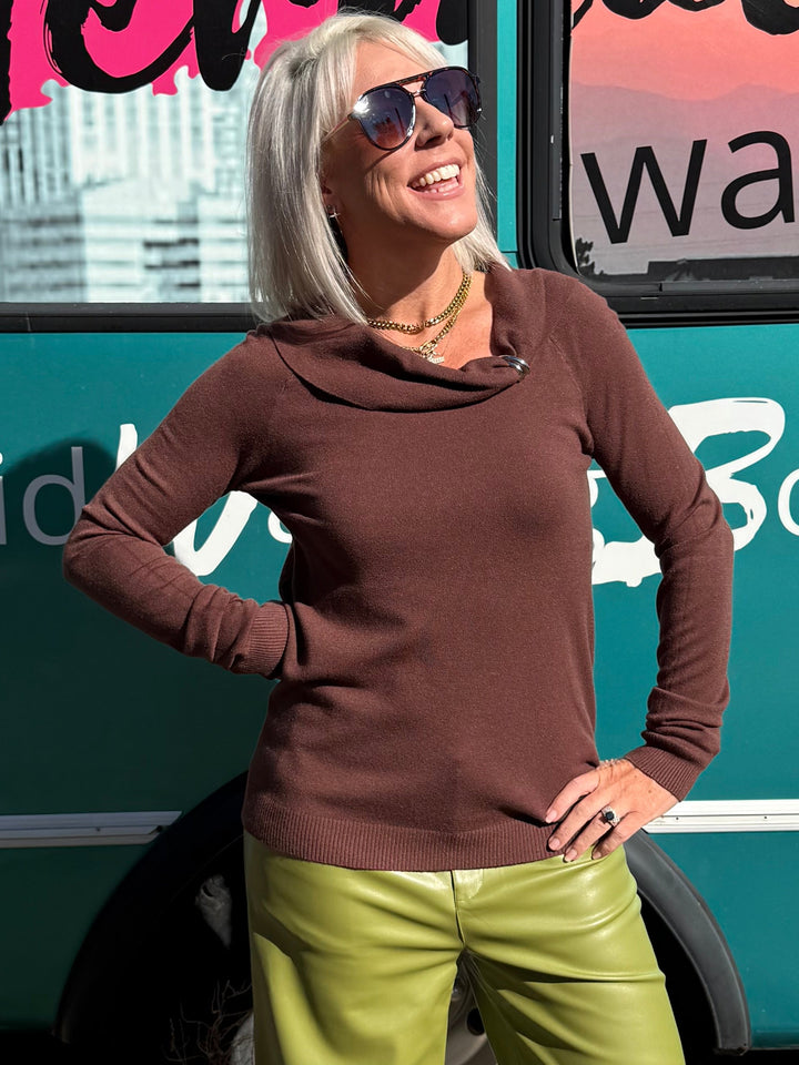 A woman wearing a brown long-sleeve cowl-neck top and olive green wide-leg pants stands in front of a teal bus with &
