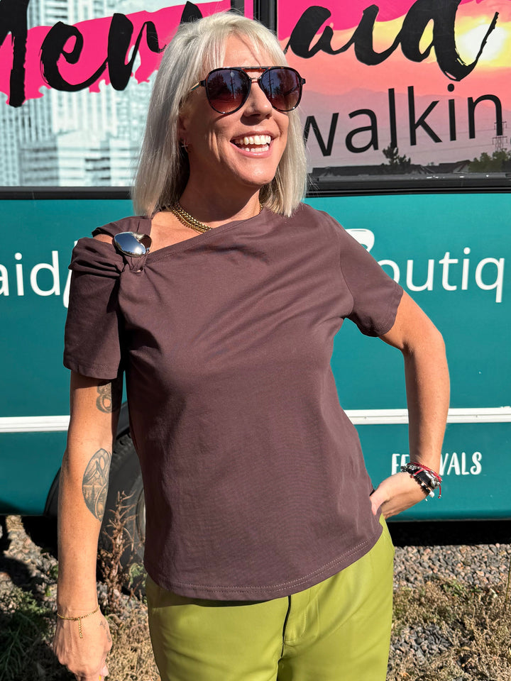 A woman with blonde hair and sunglasses stands smiling in front of a teal bus with &