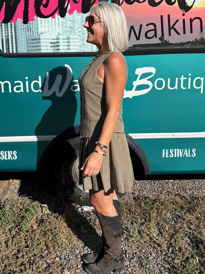 A woman with short white hair stands in profile in front of a teal bus with large windows and a pink sunset graphic. The bus has the words &