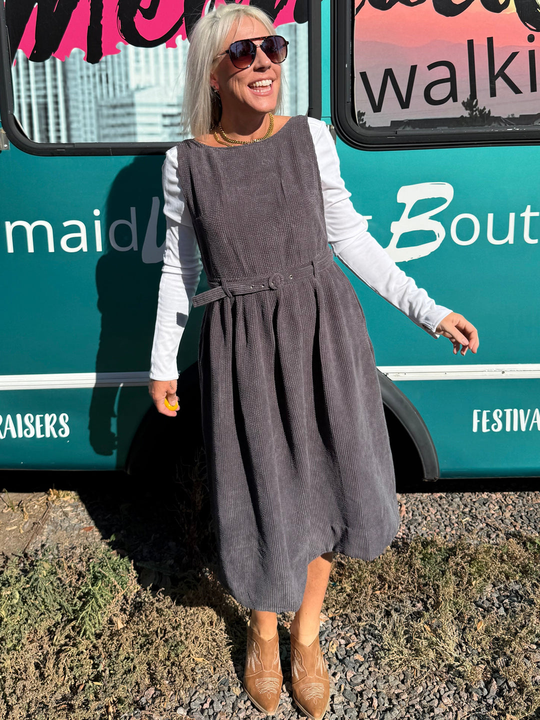 A woman wearing a dark gray pinafore dress over a white long-sleeve shirt and brown boots stands next to a teal bus with &