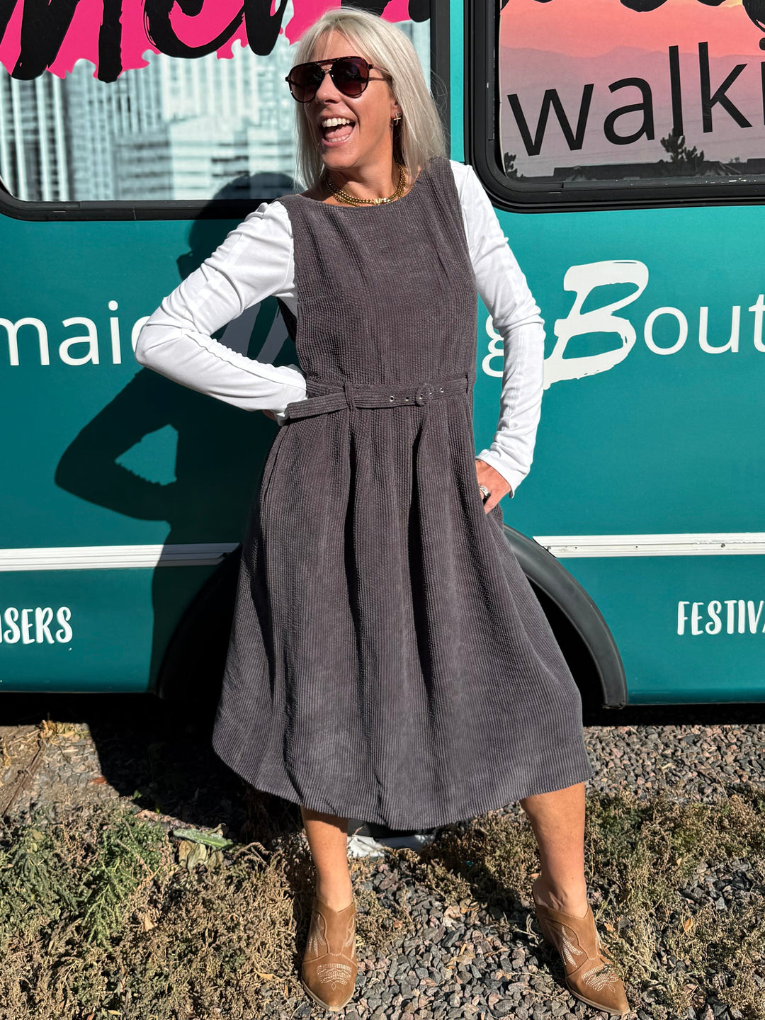 A woman wearing a dark gray sleeveless pinafore dress over a white long-sleeve shirt stands next to a teal bus with &