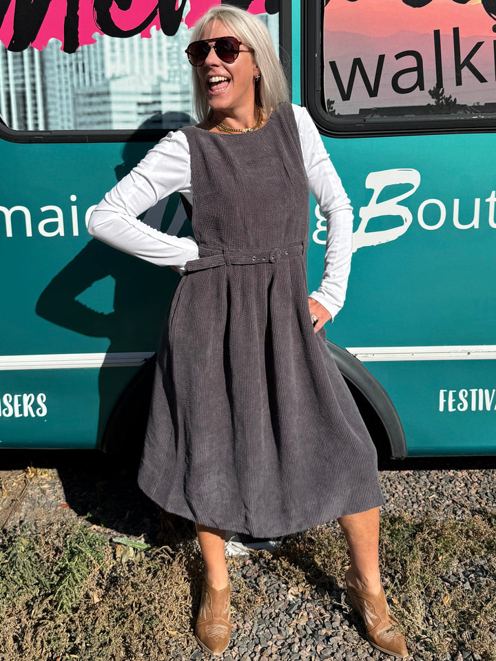 A woman wearing a dark gray sleeveless pinafore dress over a white long-sleeve shirt stands next to a teal bus with &