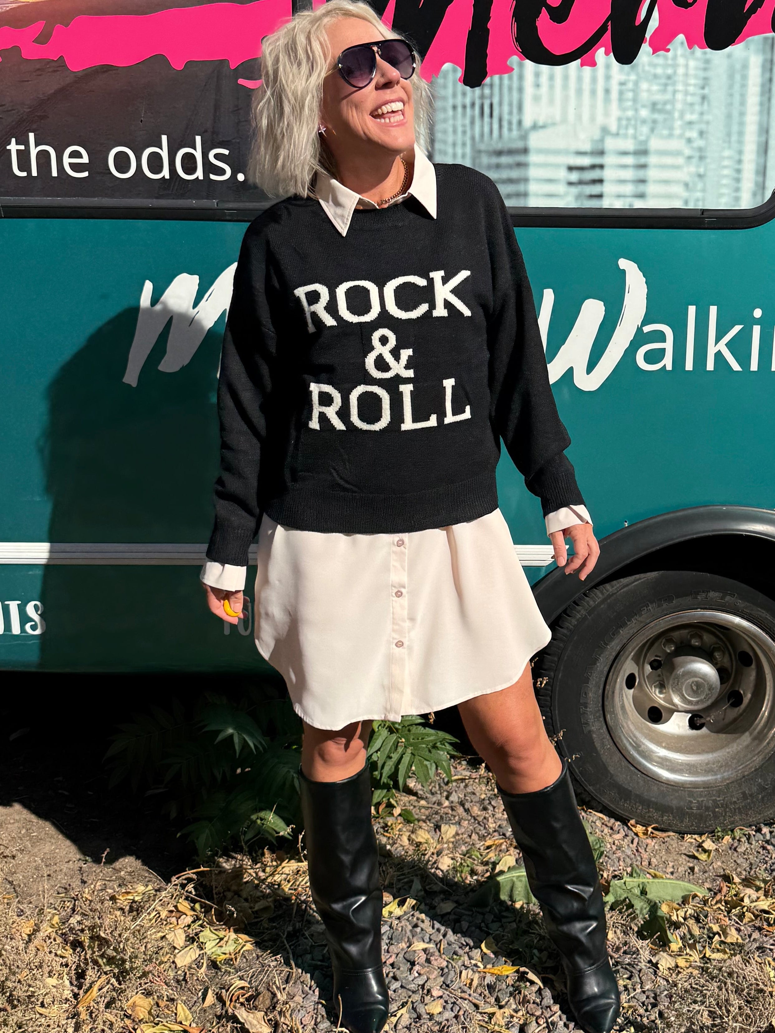 Person wearing a 'Rock & Roll' sweatshirt in front of a teal bus with reflective surfaces.