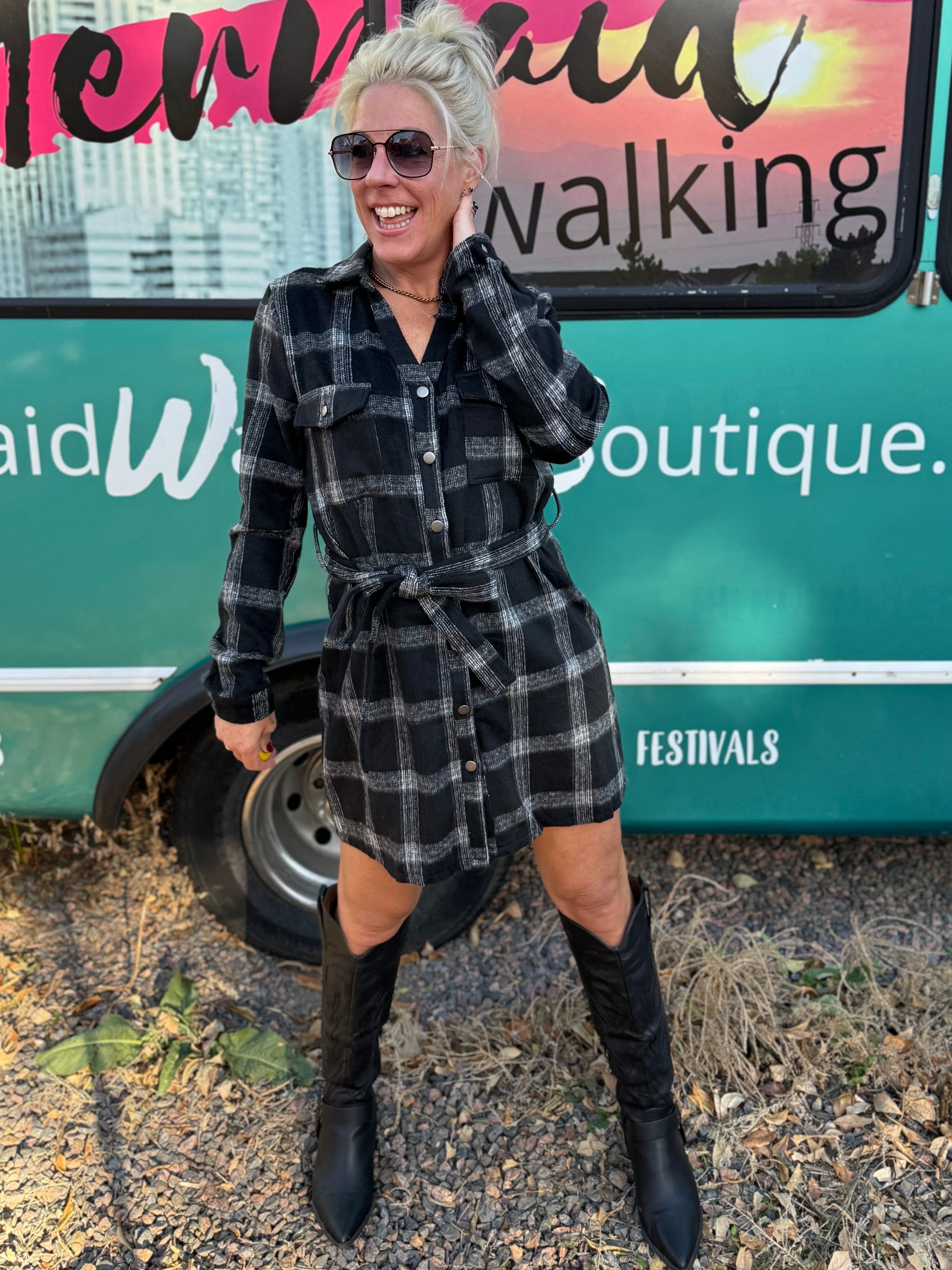 Woman in a plaid dress and boots standing in front of a mobile boutique bus.