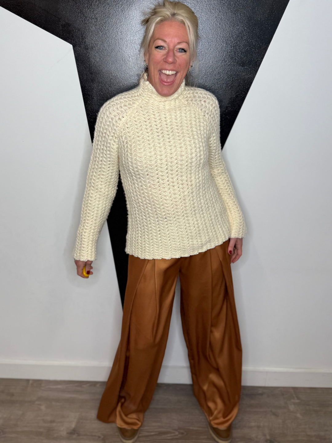 Person wearing a cream sweater and brown pants standing against a black and white geometric wall.