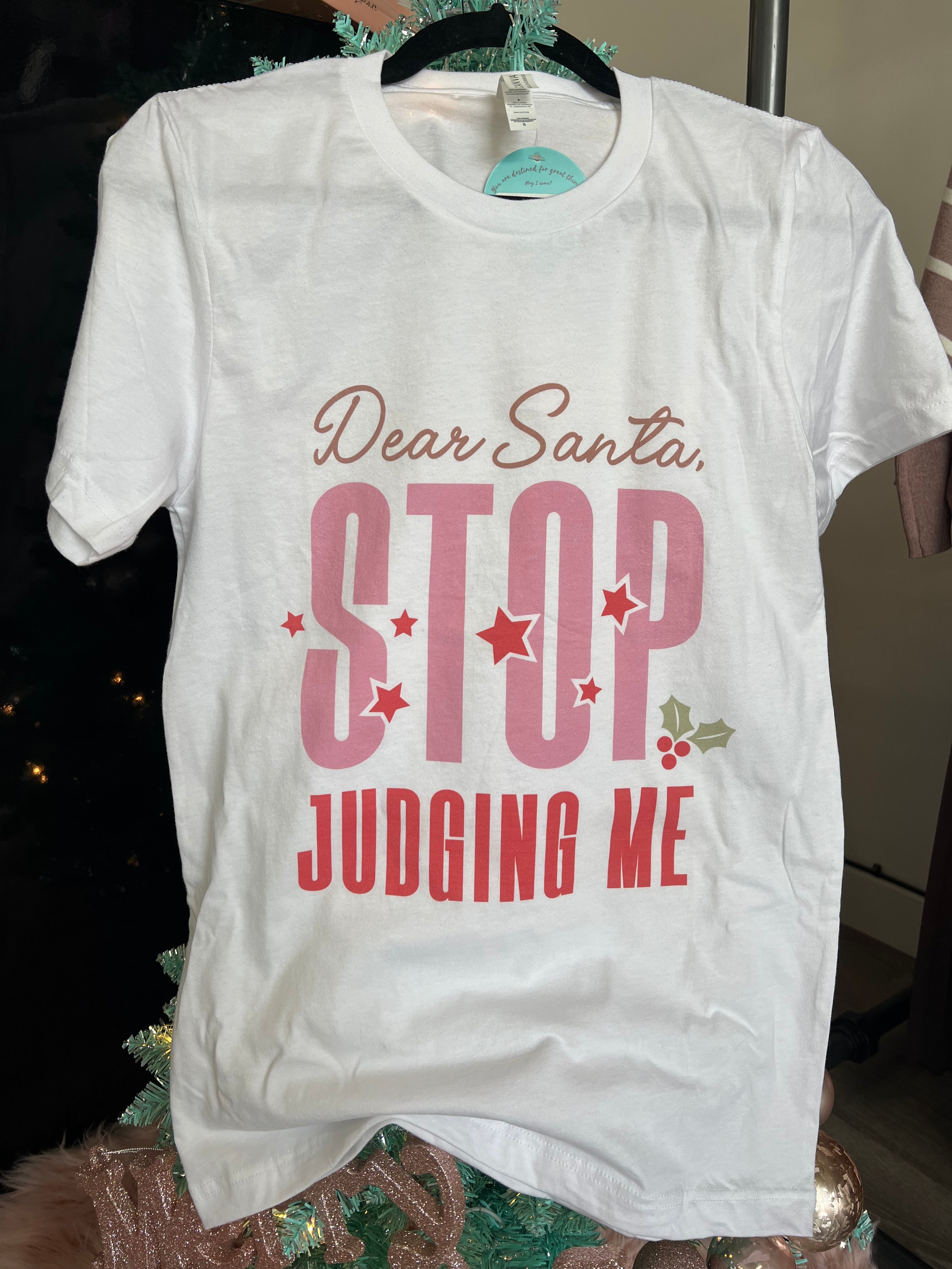 A white short-sleeved t-shirt with text graphic reading 'DEAR SANTA, STOP JUDGING ME' displayed on it, hanging on a hanger with a Christmas tree and decorations in the background.