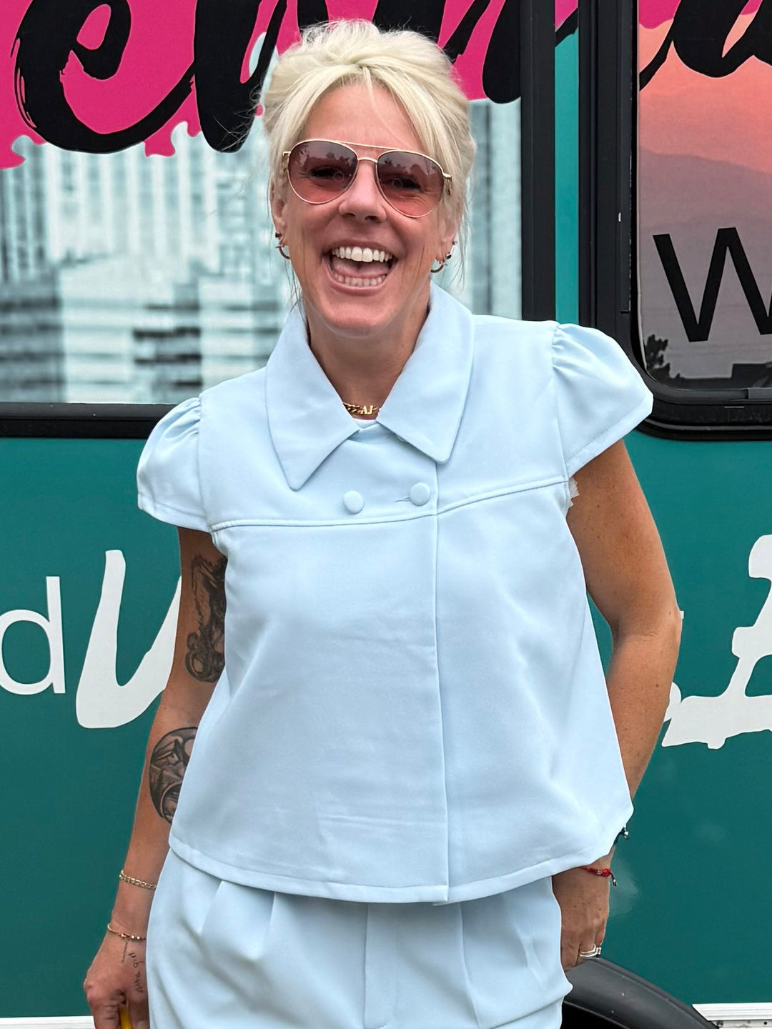 A woman wearing a light blue short-sleeve button-up shirt and matching wide-leg pants stands in front of a teal mobile boutique bus with &