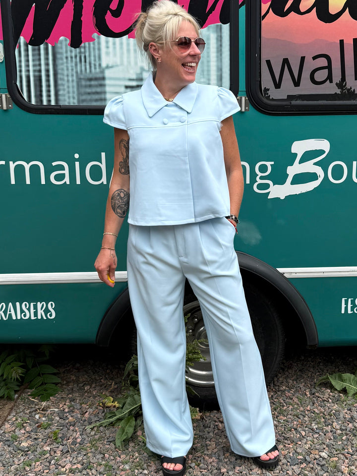 A woman stands in front of a teal bus with large windows and printed text including &