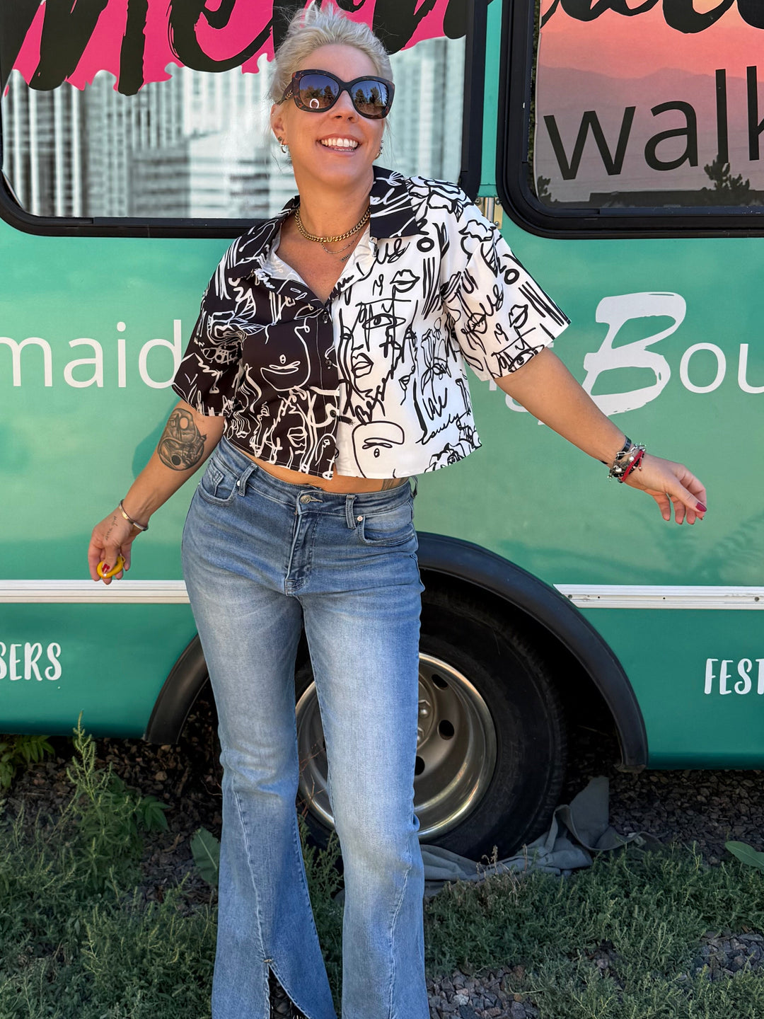A woman wearing a black and white patterned short-sleeve crop top and light blue flared jeans stands in front of a turquoise bus with &
