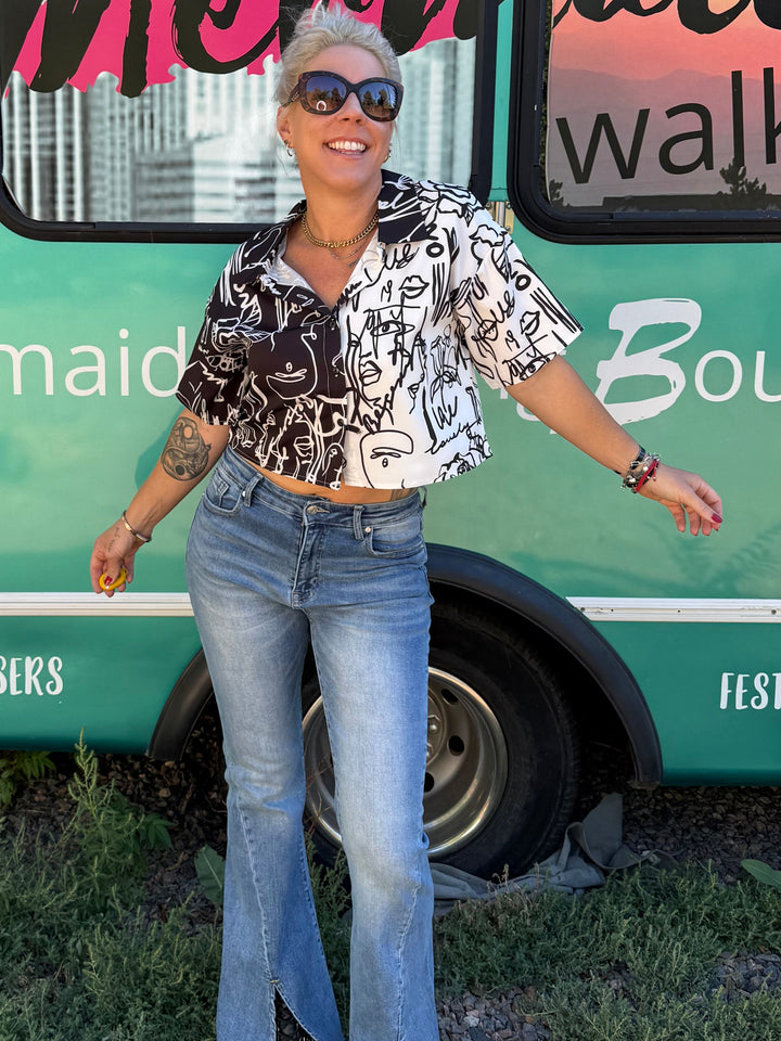 A woman wearing a black and white patterned short-sleeve crop top and light blue flared jeans stands in front of a turquoise bus with &