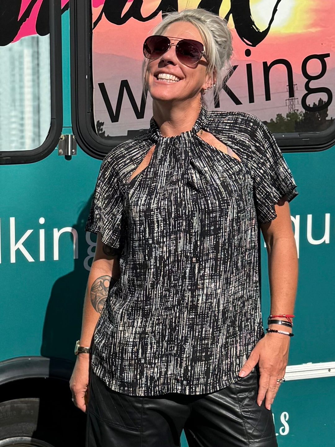 A woman wearing a black and white patterned short-sleeve top, black pants, and black sandals stands in front of a teal bus with &