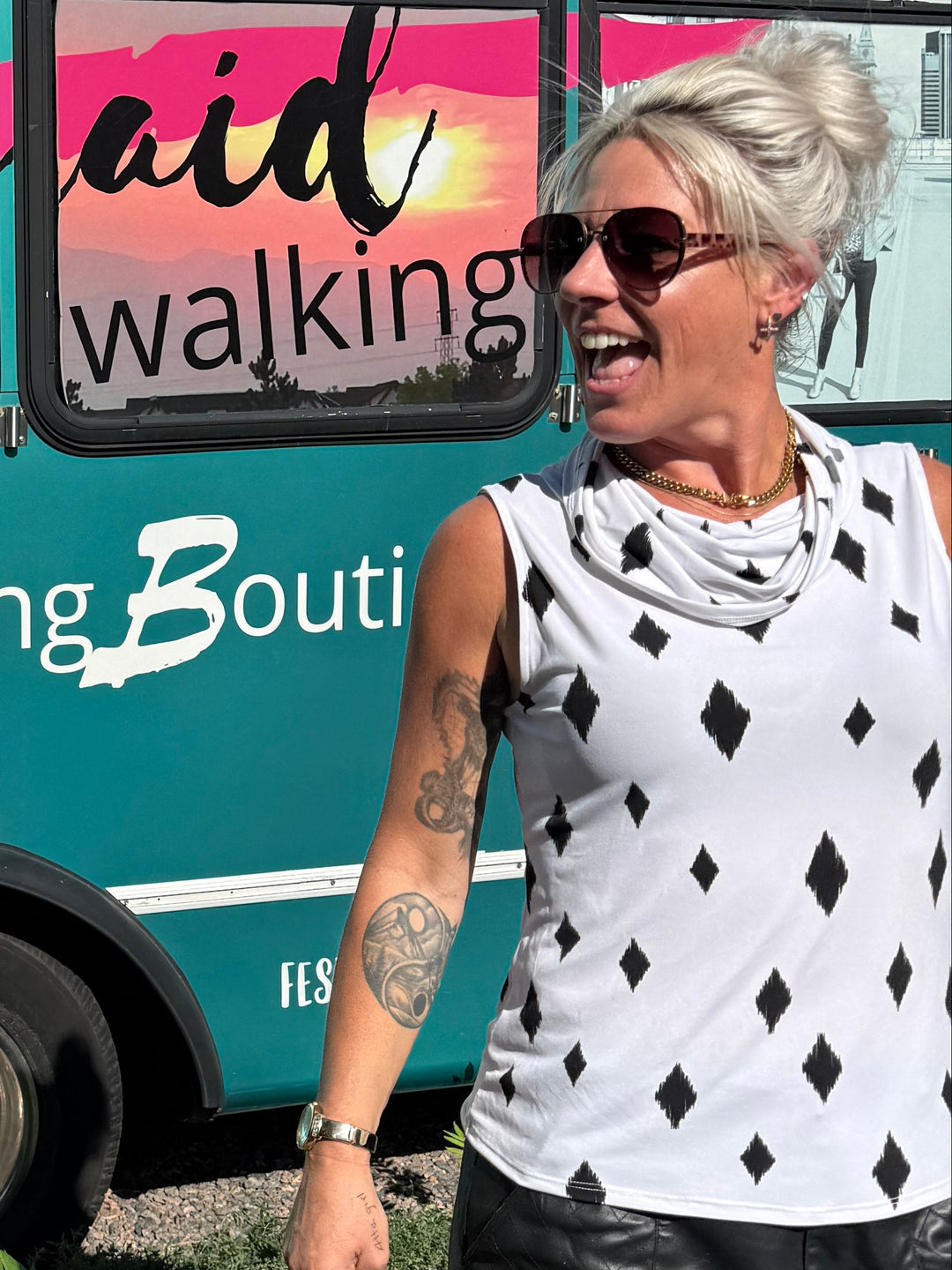 A woman with short gray hair and sunglasses stands next to a teal bus printed with the words 'Mermaid Walking Boutic' and colorful window graphics, outdoors on a sunny day.
