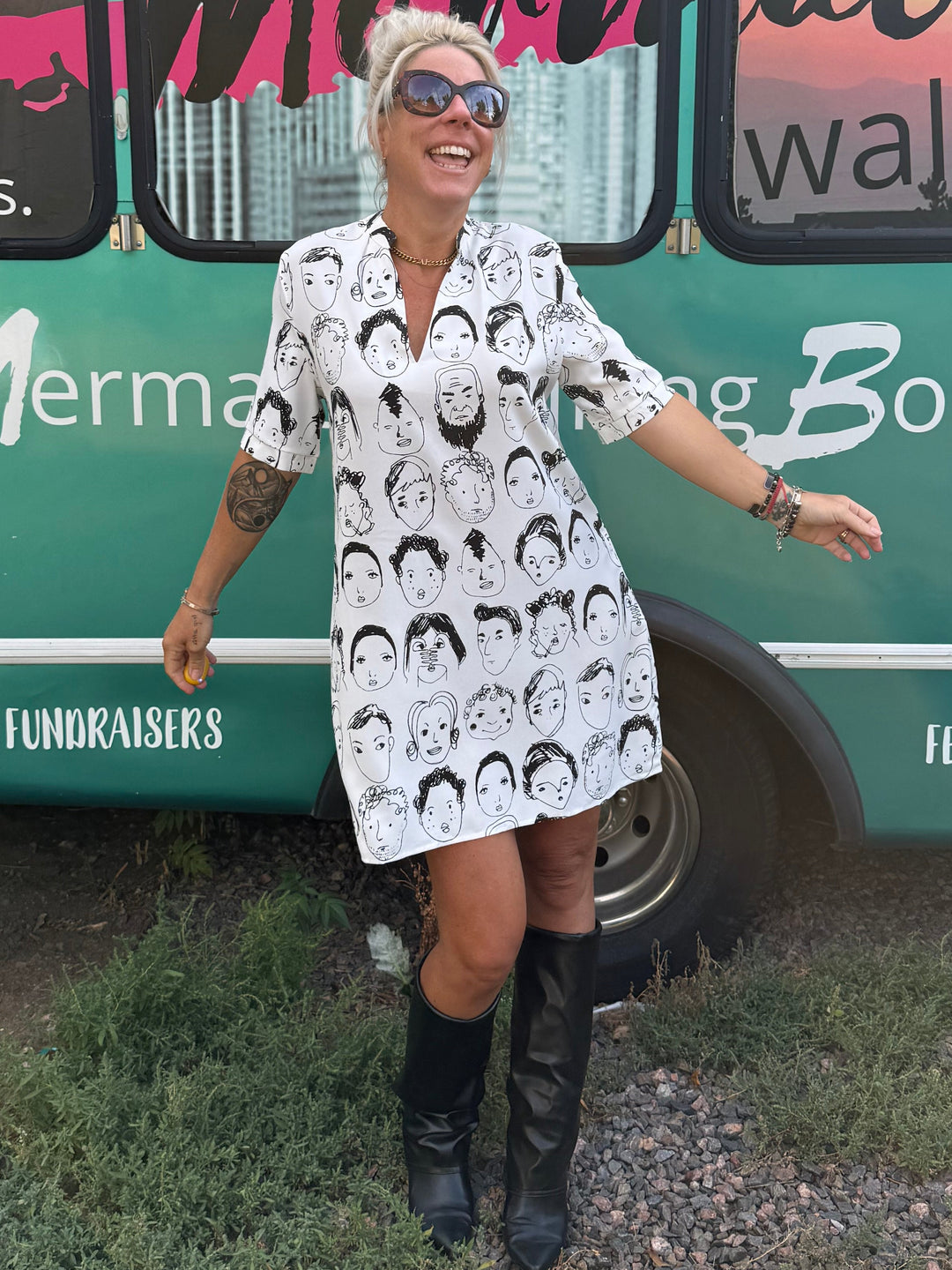 A woman wearing a white short-sleeve button-up dress with a black and white mermaid silhouette pattern stands in front of a teal bus printed with 'Mermaid Walking Boutique'.