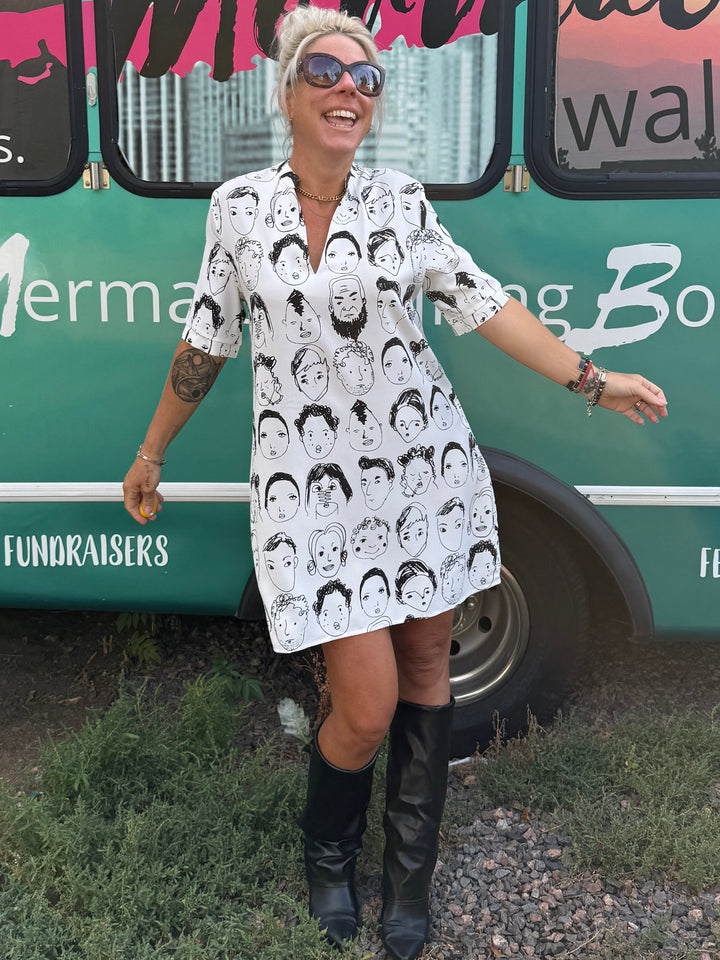 A woman wearing a white short-sleeve button-up dress with a black and white mermaid silhouette pattern stands in front of a teal bus printed with &