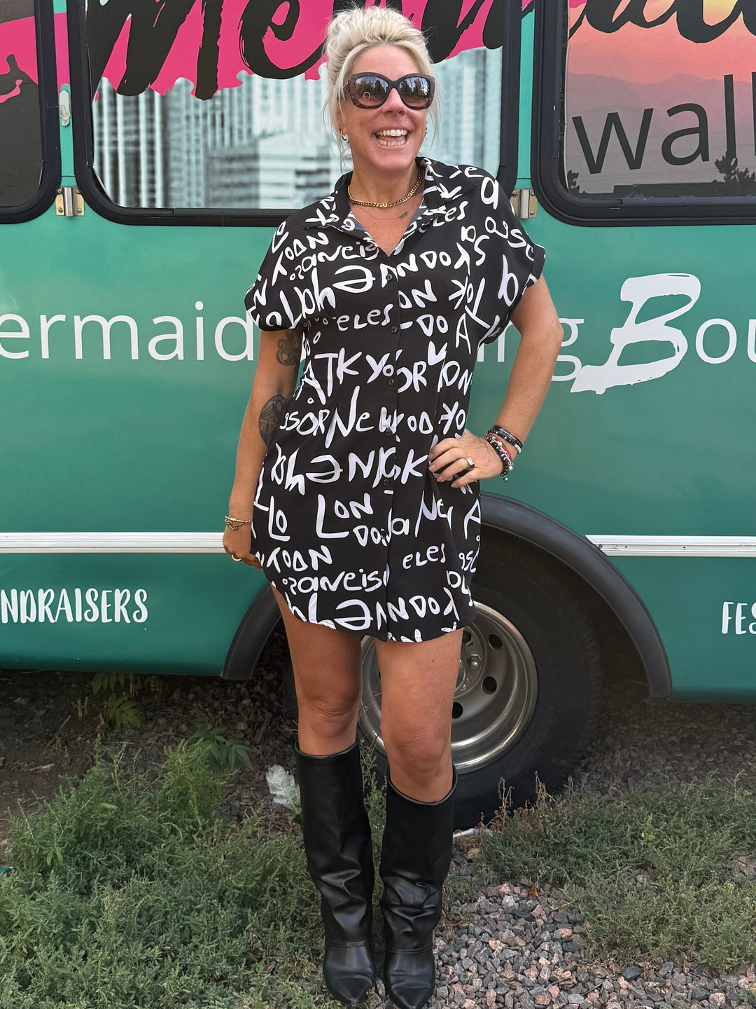 A woman stands in front of a teal bus with 'Mermaid Walking Boutique' written across the side, wearing a black-and-white patterned mini dress and black knee-high boots.