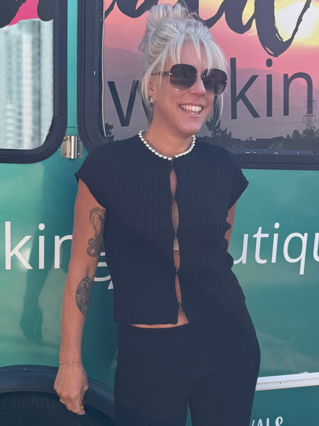 A woman wearing a black two-piece outfit with white sneakers stands in front of a teal mobile boutique bus. The bus has large windows with a sunset reflection and text including 'Mermaid Walking Boutique' and 'mermaidwalkingboutique.