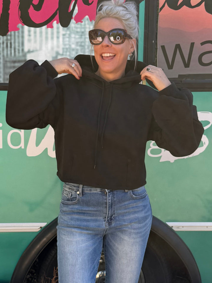 A woman wearing a black long-sleeve crop top and light blue flared jeans stands in front of a teal bus with &