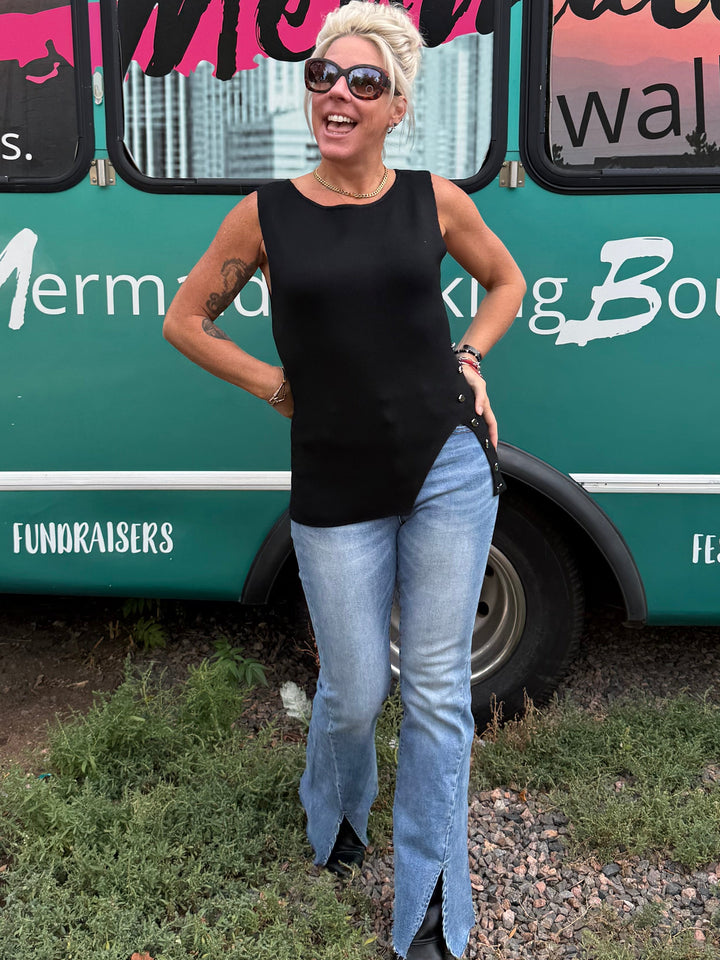 A woman wearing a black sleeveless top and light blue flared jeans stands in front of a teal event bus with &