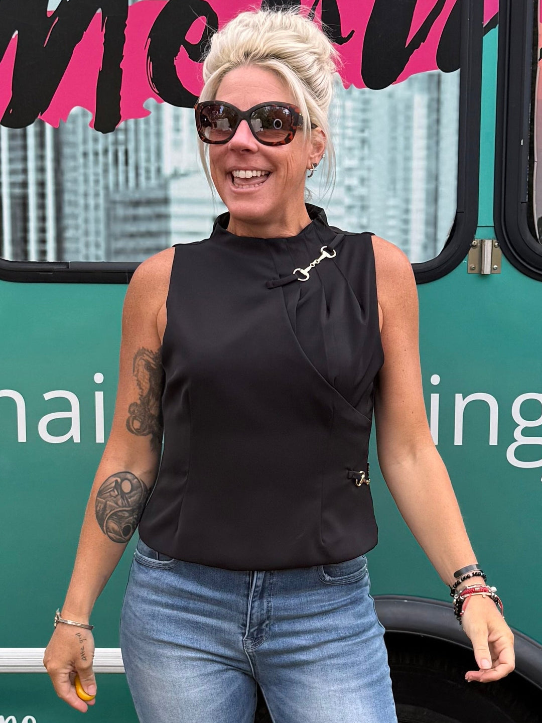 A woman wearing a black sleeveless mock-neck top and light blue flared jeans with side slits, standing in front of a green and pink 'Mermaid Fundraisers' bus.