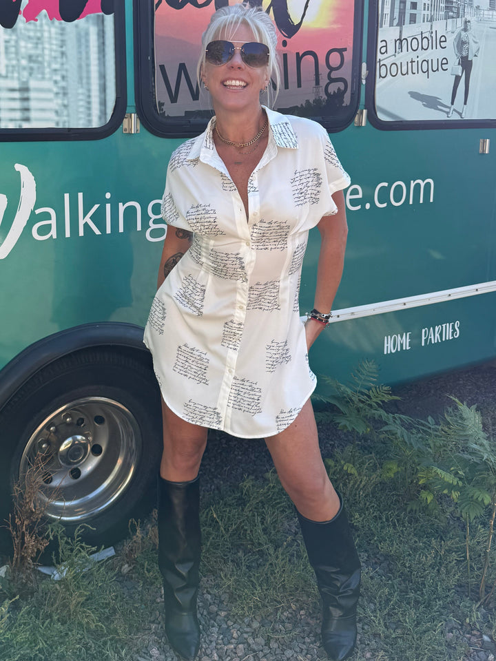 A woman wearing a cream-colored short-sleeve button-up shirt dress with a black script print, styled with black knee-high boots and sunglasses, posing in front of a green bus.