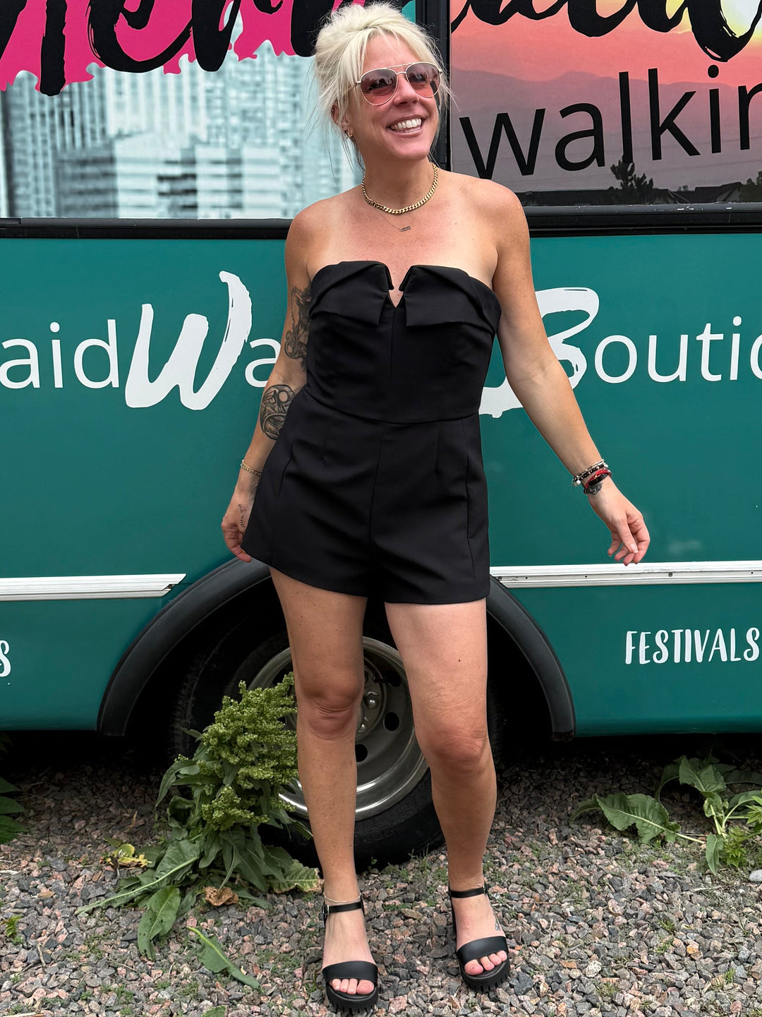 A woman stands in front of a teal bus with large windows and colorful graphics, wearing a black strapless mini dress and black sandals. The bus has visible text including &