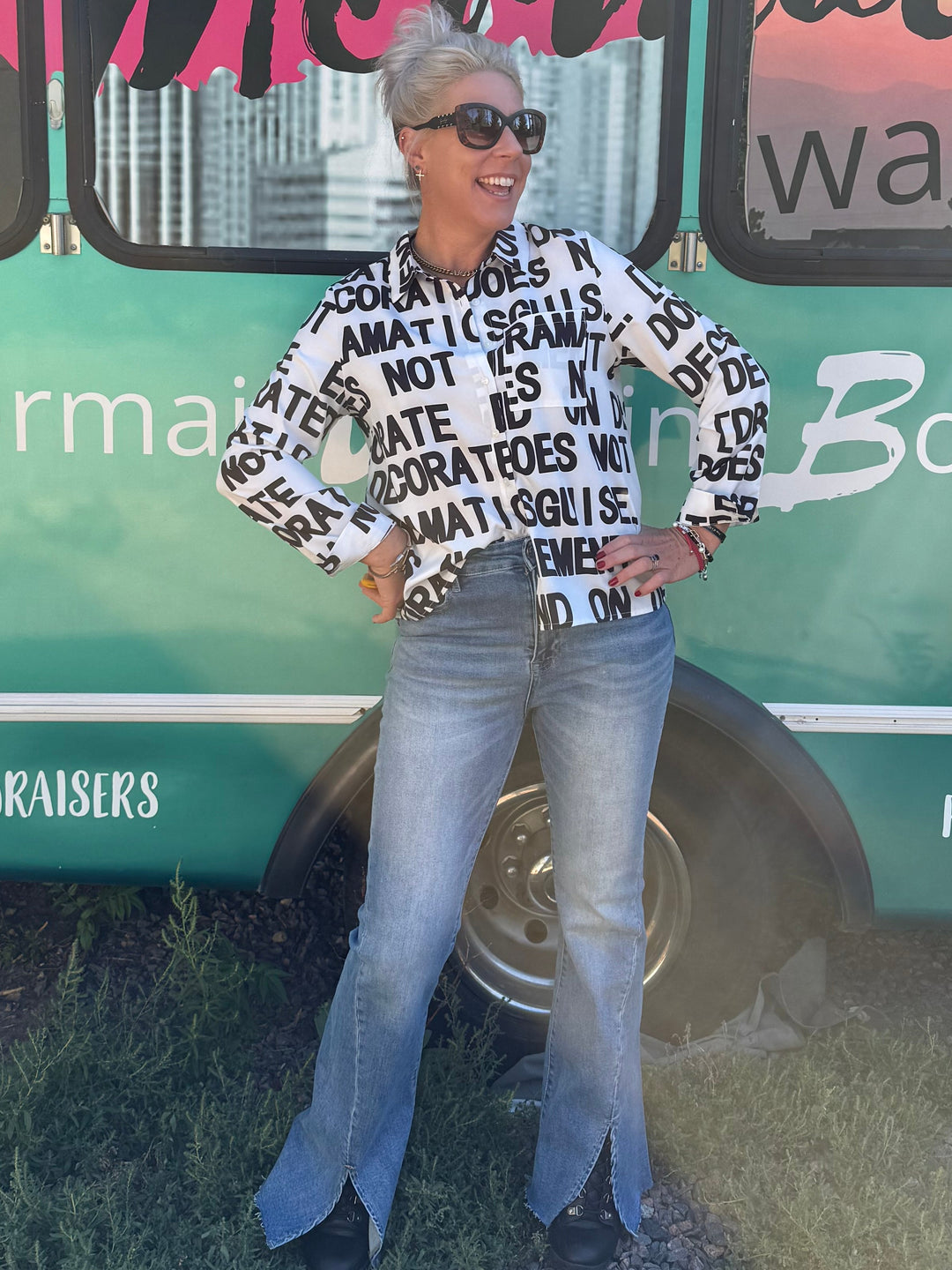 A woman wearing a black and white patterned long-sleeve top, light blue flared jeans, and sunglasses stands in front of a teal bus with 'Mermaid Walking Boutique' written on the side.