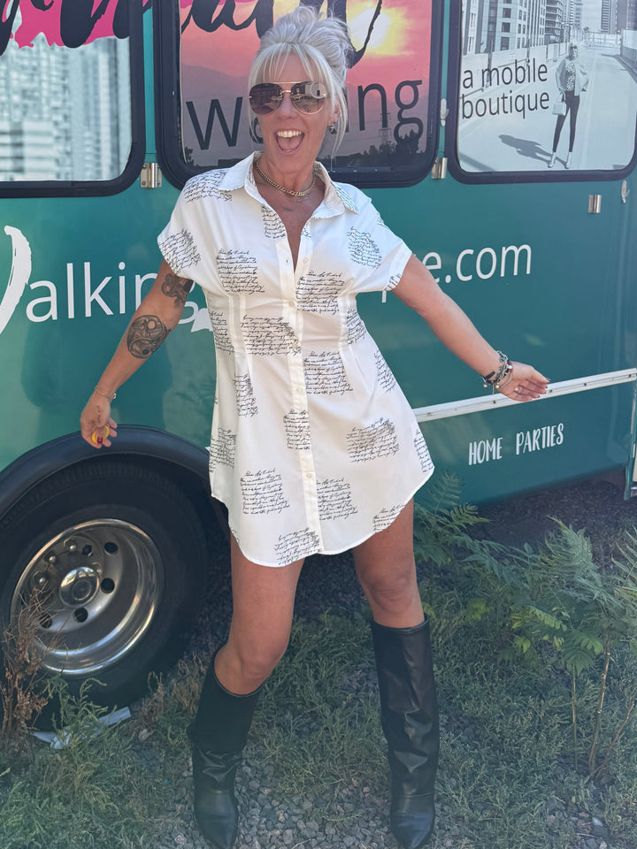 A woman with blonde hair in a bun and sunglasses stands outdoors in front of a green bus, wearing a white short-sleeve button-up shirt dress covered in black script text, paired with black knee-high boots.