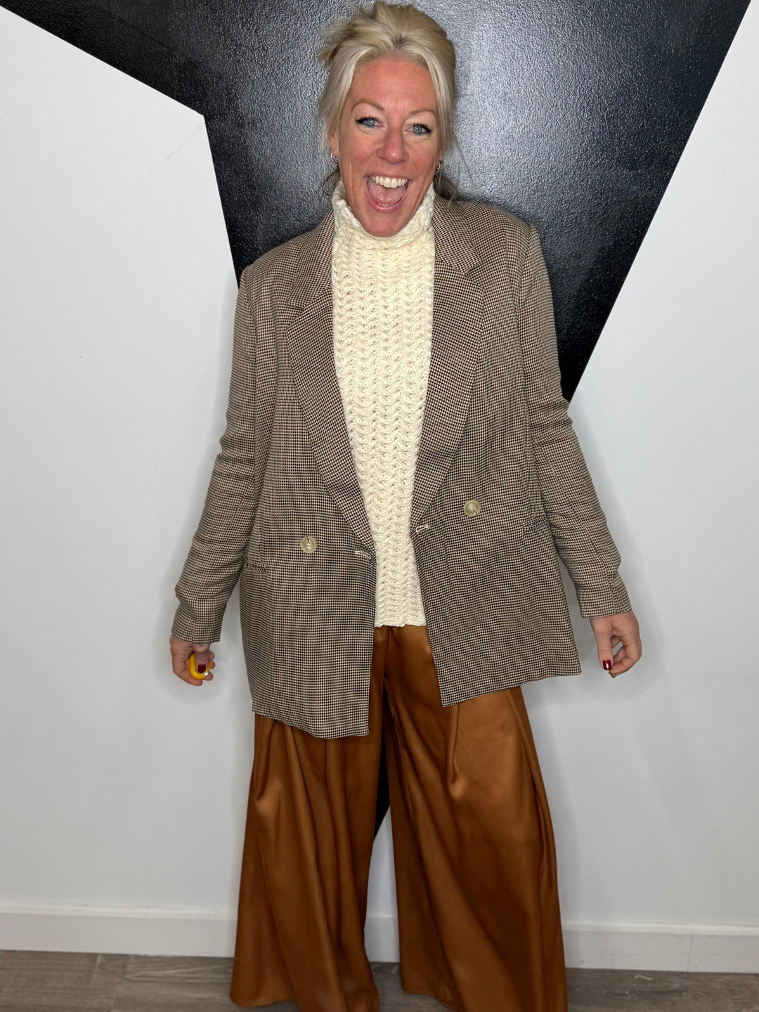 Woman wearing a beige blazer, white sweater, and brown pants against a black and white geometric wall.