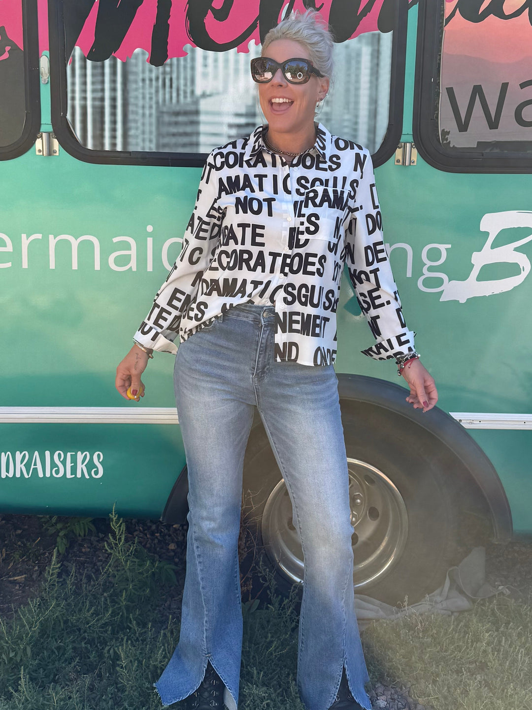 A woman wearing a black and white patterned long-sleeve top, light blue flared jeans, and black shoes stands in front of a teal bus with &