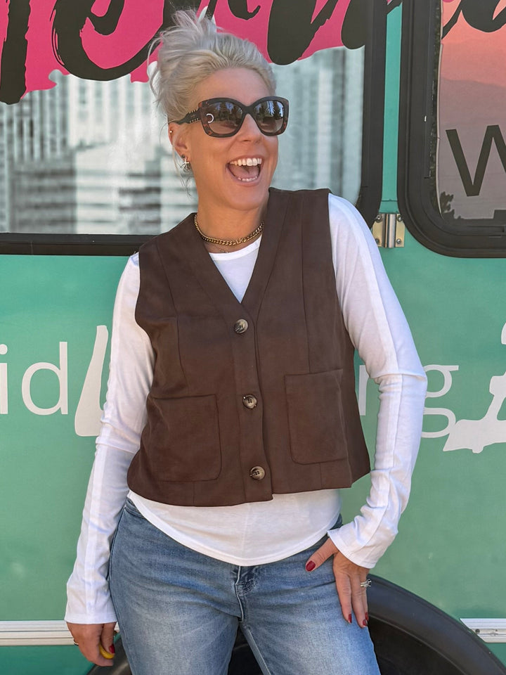 A woman wearing a brown vest, white long-sleeve shirt, and blue flared jeans stands in front of a teal bus with &