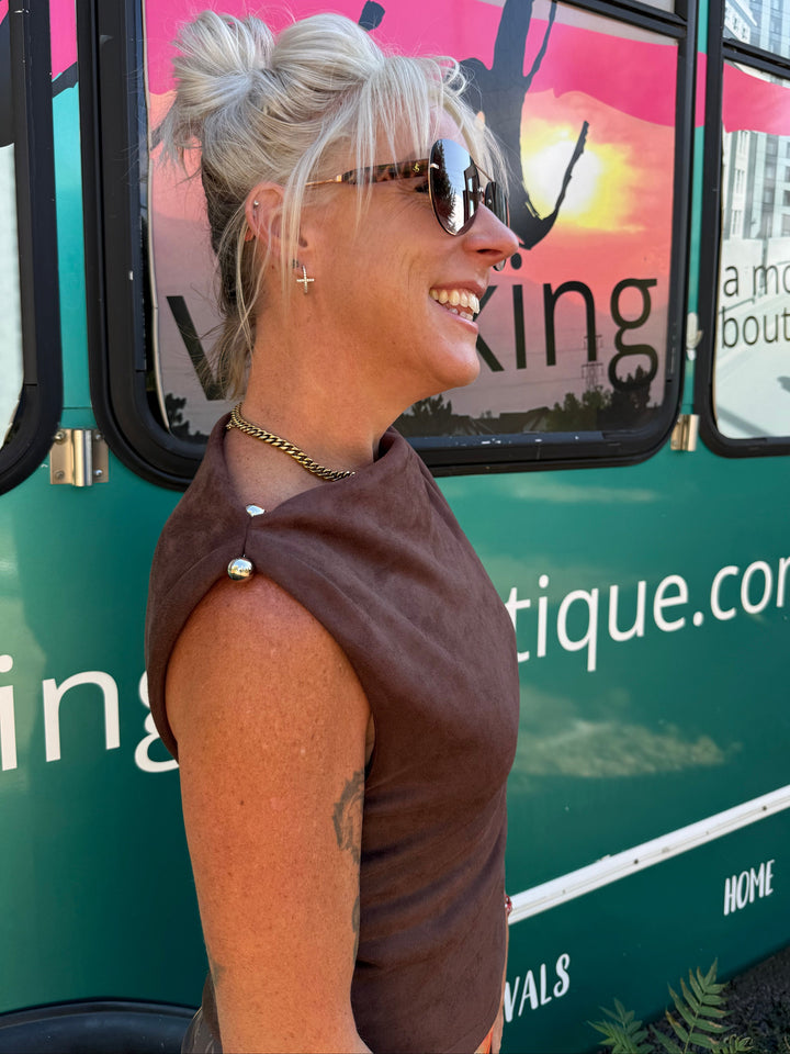 A woman with blonde hair in a bun, wearing a brown sleeveless top and sunglasses, standing in profile in front of a green mobile boutique truck.