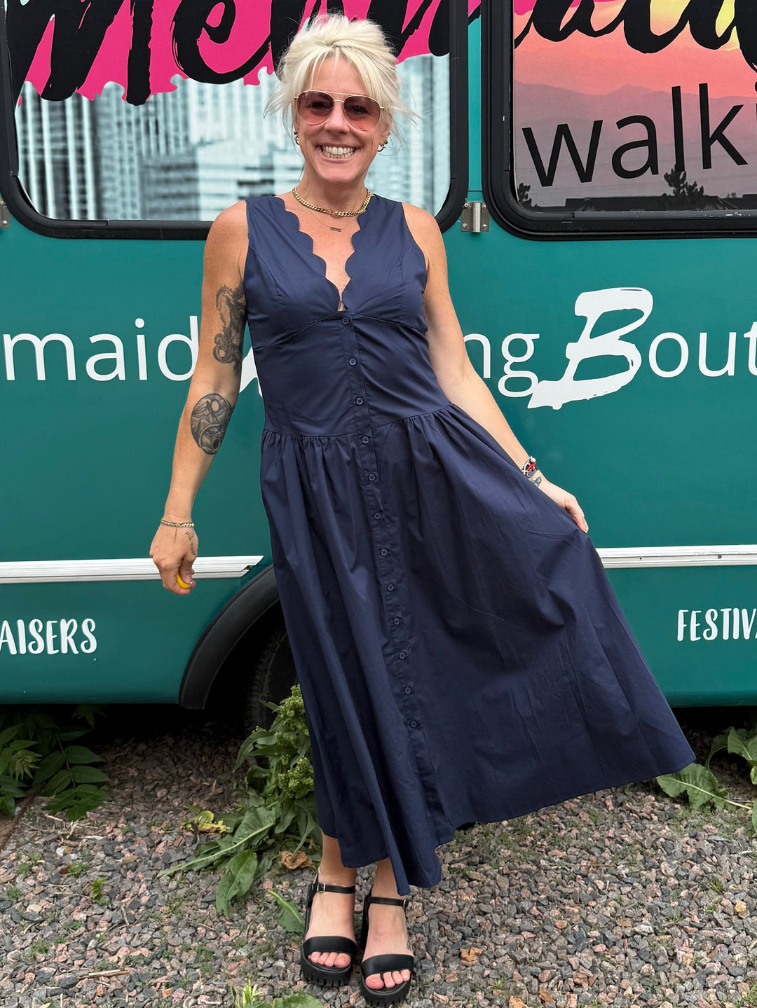 A woman wearing a navy blue sleeveless maxi dress and black sandals stands in front of a teal mobile boutique bus with &