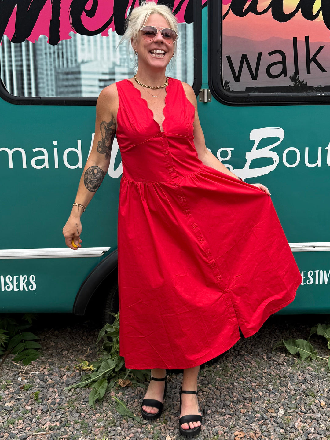 A woman wearing a bright red sleeveless maxi dress and black sandals stands in front of a teal bus with 'Mermaid Walking Boutique' written across the side windows.