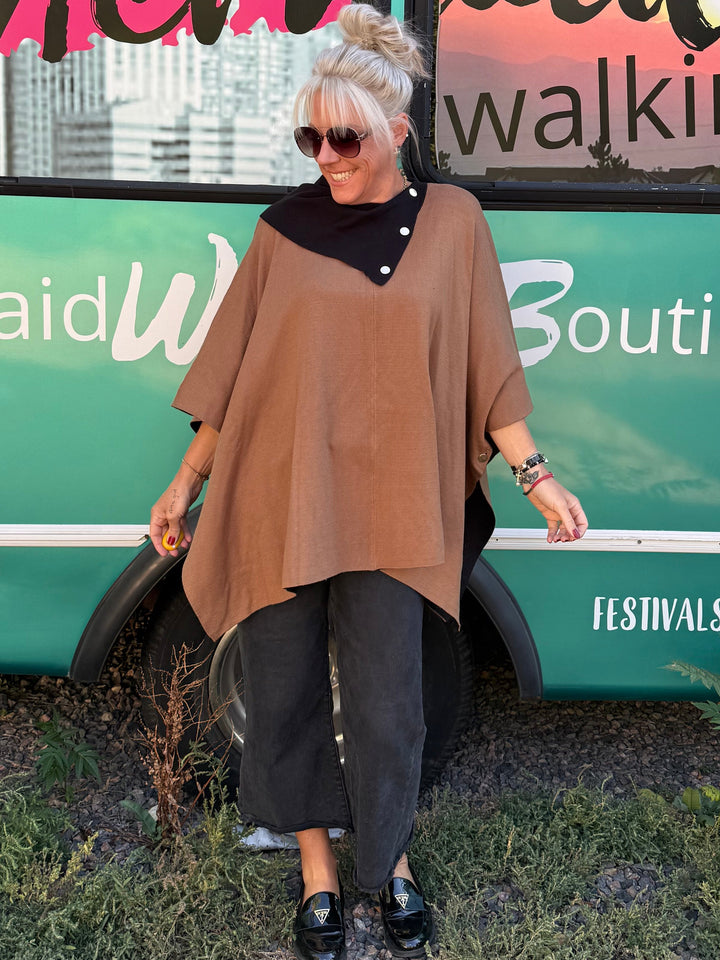 A woman wearing a brown asymmetrical poncho top, black pants, and black loafers stands in front of a turquoise bus with &