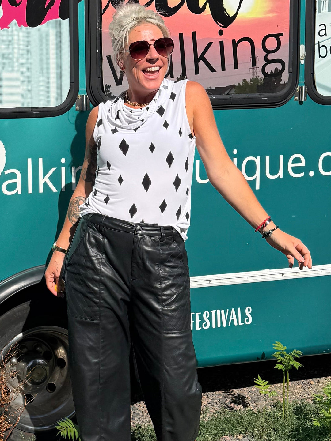 A woman wearing sunglasses, a white sleeveless top with black polka dots, and black pants stands next to a teal Mermaid Walking Boutique vehicle outdoors.