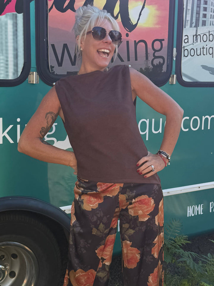A woman posing in front of a teal mobile boutique bus. She is wearing a sleeveless brown top and wide-leg brown pants with a red and orange floral pattern.