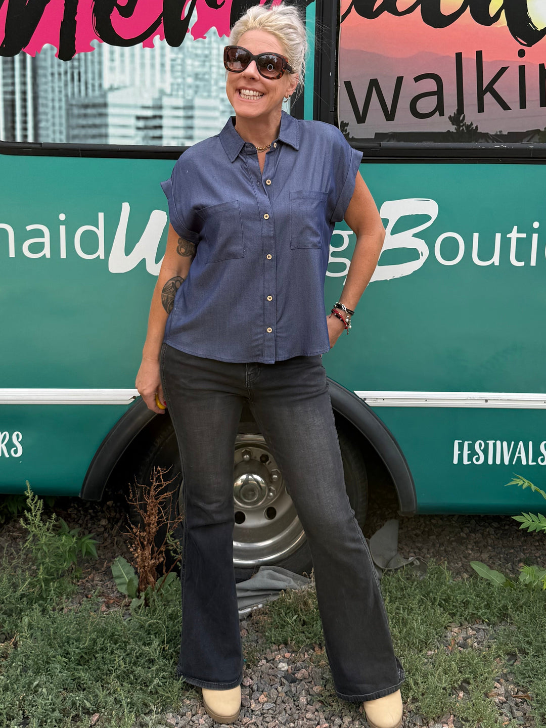A woman wearing a blue button-down shirt and dark jeans stands in front of a teal bus with 'Mermaid Walking Boutique' written on the side.