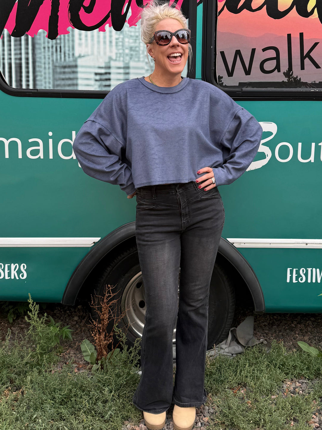 A woman wearing a slate blue long-sleeve sweatshirt and black flared jeans stands in front of a teal bus with &