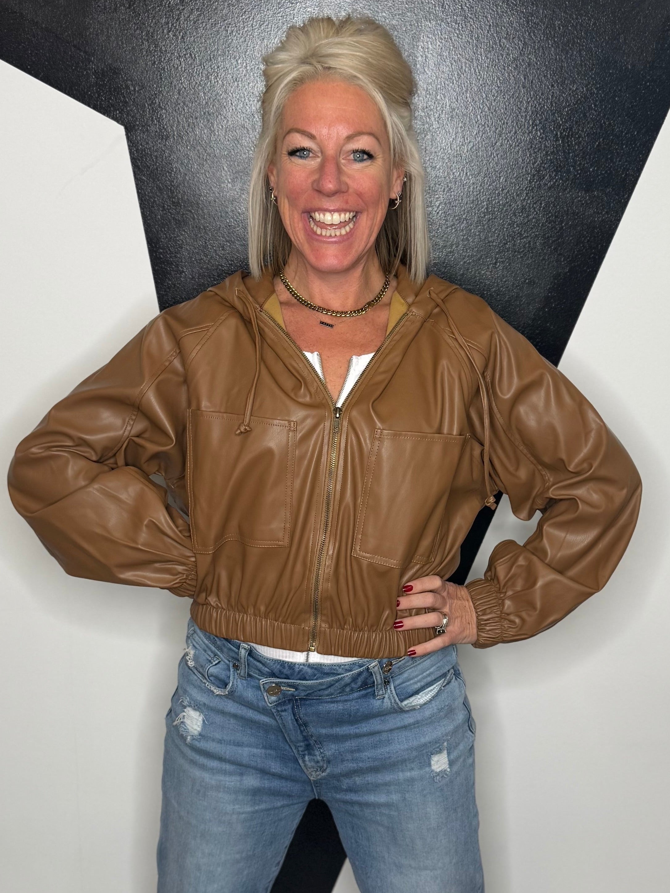 Woman wearing a brown leather jacket and blue jeans standing against a black and white geometric wall.