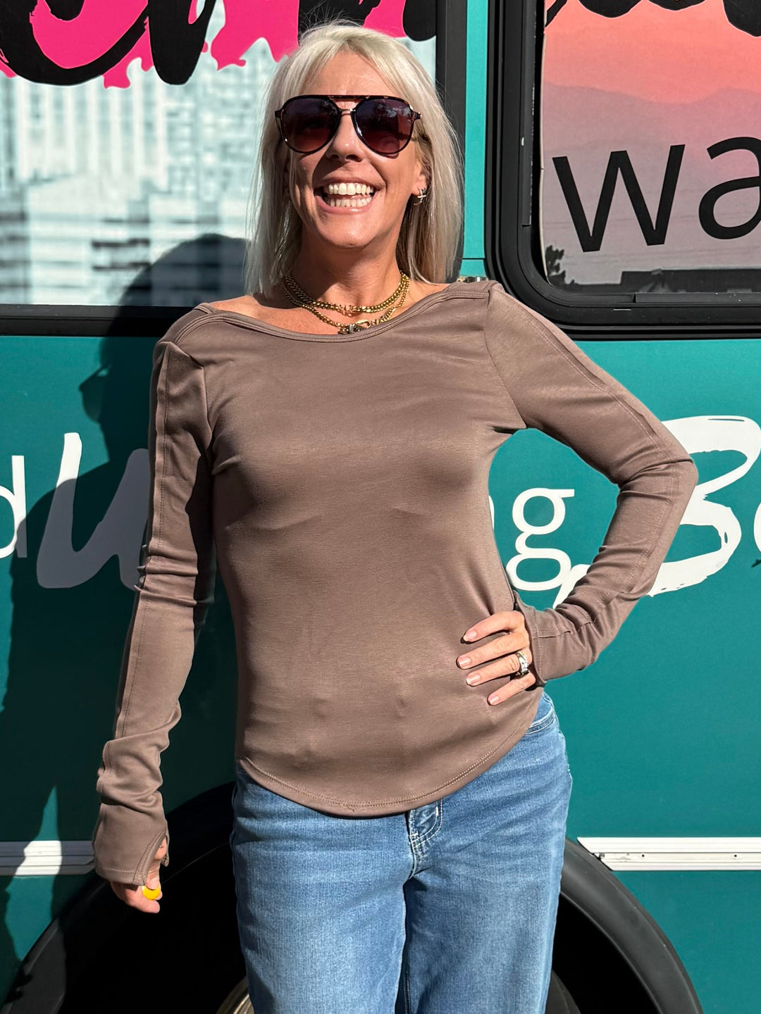 A woman wearing a brown long-sleeve top and blue jeans stands in front of a teal bus with &