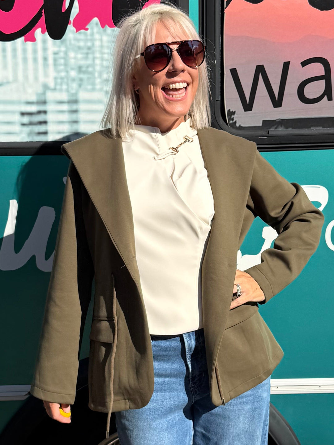 A woman with white hair and sunglasses stands in front of a teal bus with &