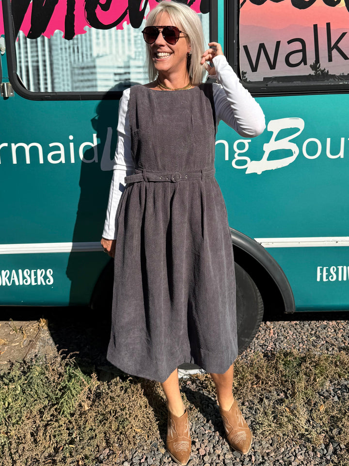 A woman wearing a dark gray corduroy pinafore dress over a white long-sleeve shirt stands in front of a teal bus with &