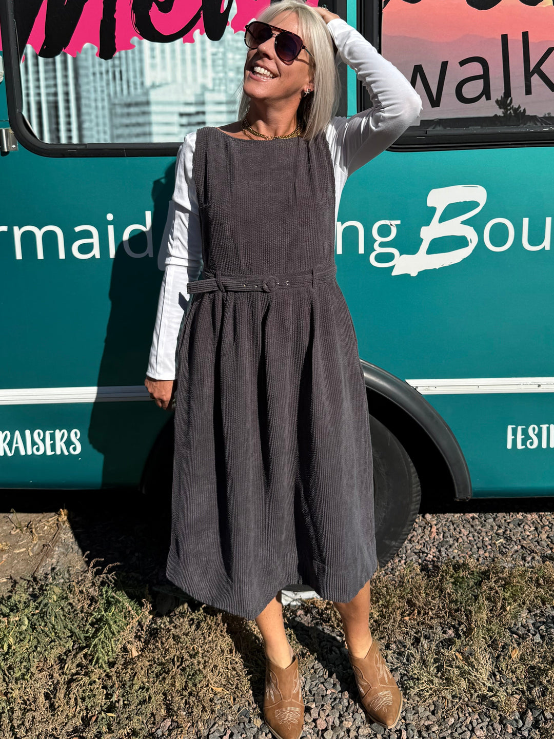 A woman wearing a dark gray pinafore dress over a white long-sleeve shirt stands in front of a teal bus with &